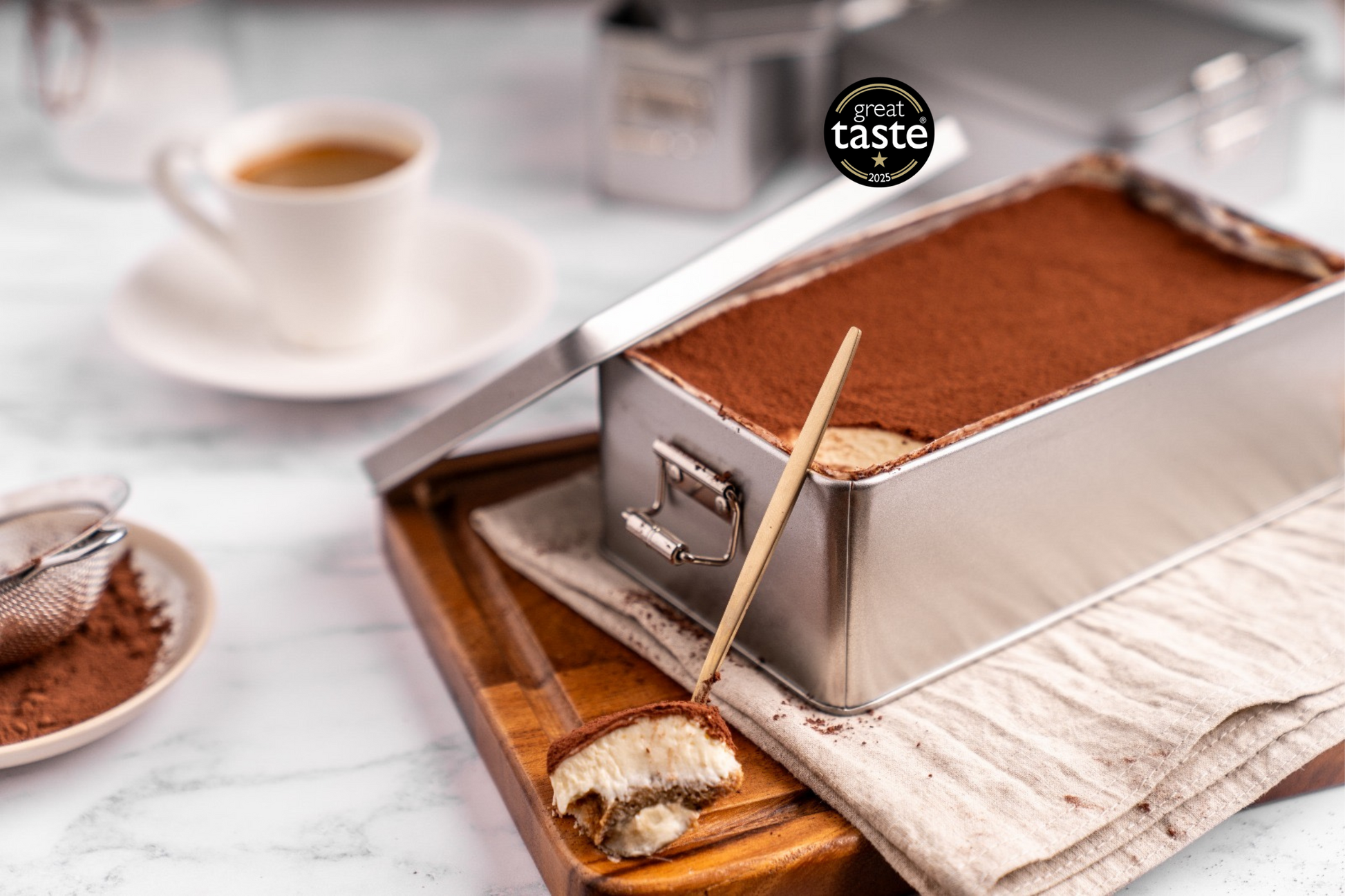 Classic award-winning tiramisu in a metal tin, dusted with cocoa powder, with a spoonful on a wooden board and a coffee cup in the background – Great Taste Star winner, alcohol-free Italian dessert by Tiramisu Cake UK.